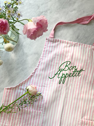 Pink and white striped apron with 'Bon Appetit' text, placed on a marble surface with flowers.