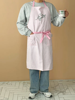 Person wearing a striped apron with 'Bon Appetit' text, holding a mug against a beige wall.