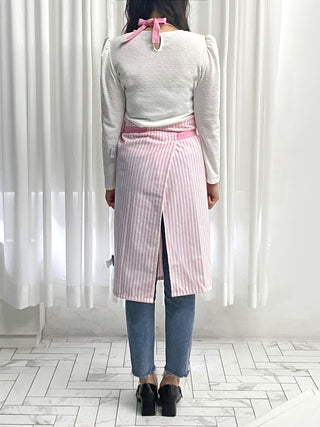 Person wearing a pink striped apron over a white shirt and blue jeans, standing in front of a white curtain.